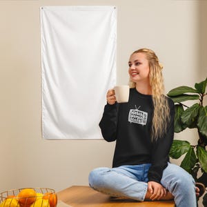 May include: A woman wearing a black sweatshirt with a white graphic of a television screen with the text "Watch Women Score" on it. She is sitting on a wooden surface and holding a white mug.
