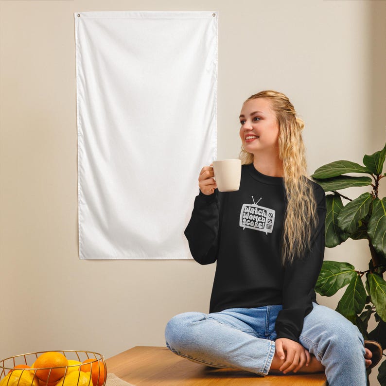 May include: A woman wearing a black sweatshirt with a white graphic of a television screen with the text "Watch Women Score" on it. She is sitting on a wooden surface and holding a white mug.