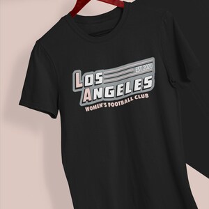Pu&ograve; includere: T-shirt nero con la scritta "LOS ANGELES WOMEN'S FOOTBALL CLUB" in bianco, rosa e grigio. La maglietta ha un design retr&ograve; con l'anno di fondazione "EST. 2020". La maglietta &egrave; appesa a una gruccia di legno.