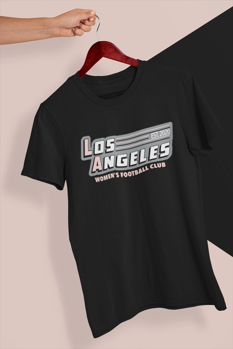 Pu&ograve; includere: T-shirt nero con la scritta "LOS ANGELES WOMEN'S FOOTBALL CLUB" in bianco, rosa e grigio. La maglietta ha un design retr&ograve; con l'anno di fondazione "EST. 2020". La maglietta &egrave; appesa a una gruccia di legno.