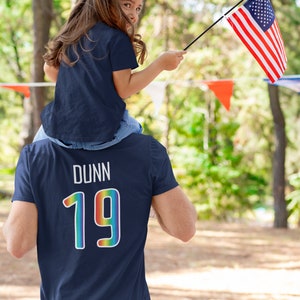 May include: A father and daughter wearing matching navy blue t-shirts. The father's shirt has the name "DUNN" and the number "19" in rainbow colors on the back. The daughter is holding a small American flag.