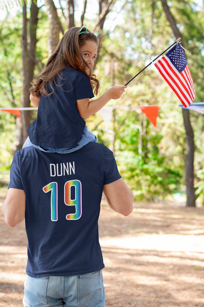 May include: A father and daughter wearing matching navy blue t-shirts. The father's shirt has the name "DUNN" and the number "19" in rainbow colors on the back. The daughter is holding a small American flag.