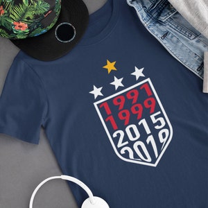 Puede incluir: Una camiseta azul marino con un dise&ntilde;o de escudo blanco que presenta los n&uacute;meros rojos 1991, 1999, 2015 y 2019. Cuatro estrellas blancas y una estrella amarilla est&aacute;n por encima del escudo.