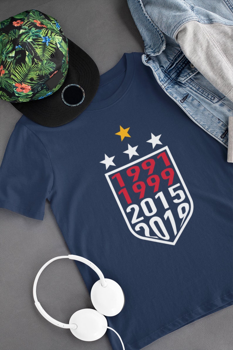 Puede incluir: Una camiseta azul marino con un dise&ntilde;o de escudo blanco que presenta los n&uacute;meros rojos 1991, 1999, 2015 y 2019. Cuatro estrellas blancas y una estrella amarilla est&aacute;n por encima del escudo.