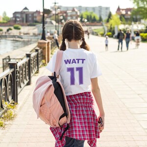May include: A young person wearing a white t-shirt with the text "WATT 11" in purple lettering. They are wearing a red and black plaid shirt over the t-shirt and carrying a pink backpack.