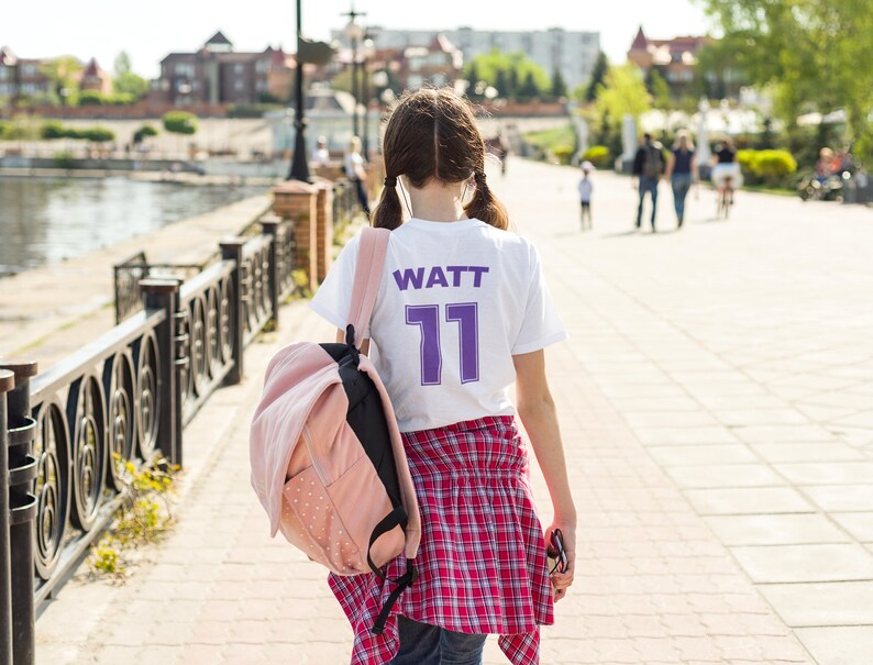 May include: A young person wearing a white t-shirt with the text "WATT 11" in purple lettering. They are wearing a red and black plaid shirt over the t-shirt and carrying a pink backpack.