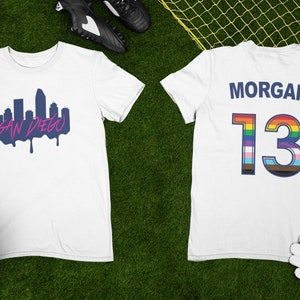 May include: White soccer jersey with a blue and pink San Diego skyline graphic on the front and the name "MORGAN" and the number "13" with a rainbow and transgender pride flag design on the back.