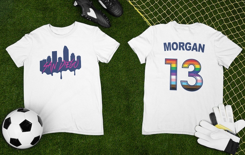 May include: White football shirt with a blue and pink San Diego skyline graphic on the front and the name "MORGAN" and the number "13" with a rainbow and transgender pride flag design on the back.