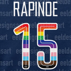 Puede incluir: Una camiseta azul con el nombre "RAPINOE" y el n&uacute;mero "15" en colores arco&iacute;ris. El n&uacute;mero est&aacute; contorneado en blanco y tiene una peque&ntilde;a bandera de EE. UU. en la parte inferior.
