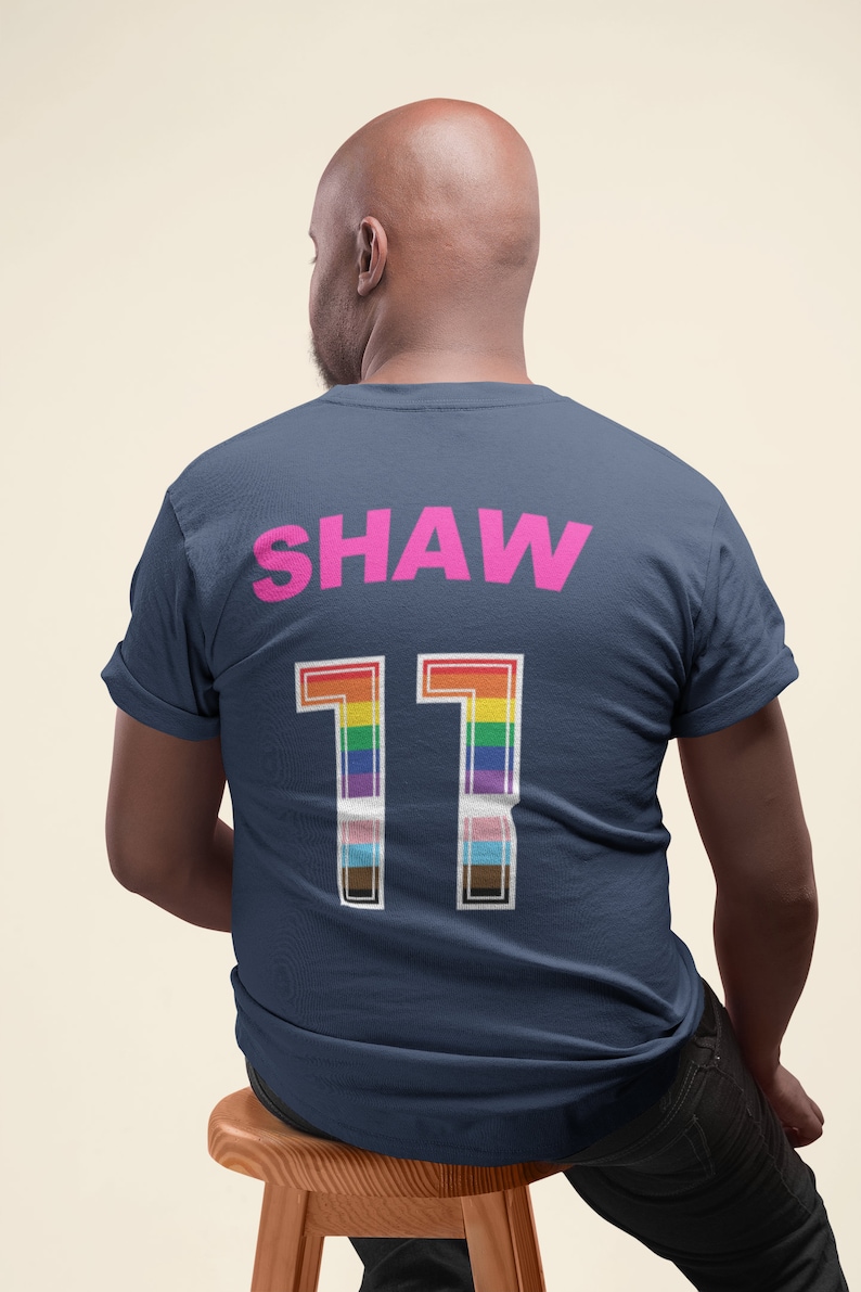 May include: A navy blue t-shirt with the name "SHAW" and the number "11" printed on the back. The number is in white with a rainbow stripe design.