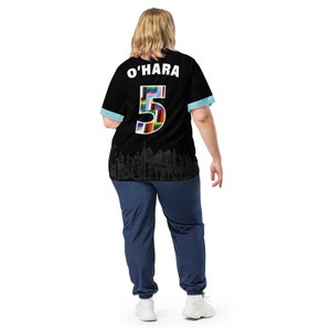 K&ouml;nnte beinhalten: Ein schwarzes Fu&szlig;balltrikot mit blauem Besatz und einer wei&szlig;en Nummer 5 auf dem R&uuml;cken. Die Nummer besteht aus kleinen Flaggen verschiedener L&auml;nder. Das Trikot tr&auml;gt auch den Namen "O'HARA" in wei&szlig; auf dem R&uuml;cken. Die Person, die das Trikot tr&auml;gt, steht mit dem R&uuml;cken zur Kamera.