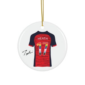 Op de afbeelding: Een ronde, witte keramische ornament met een voetbalshirtontwerp. Het rode shirt heeft marineblauwe mouwen en het regenbooggekleurde nummer 17. De naam "HEATH" staat boven het nummer, met een handtekening links.
