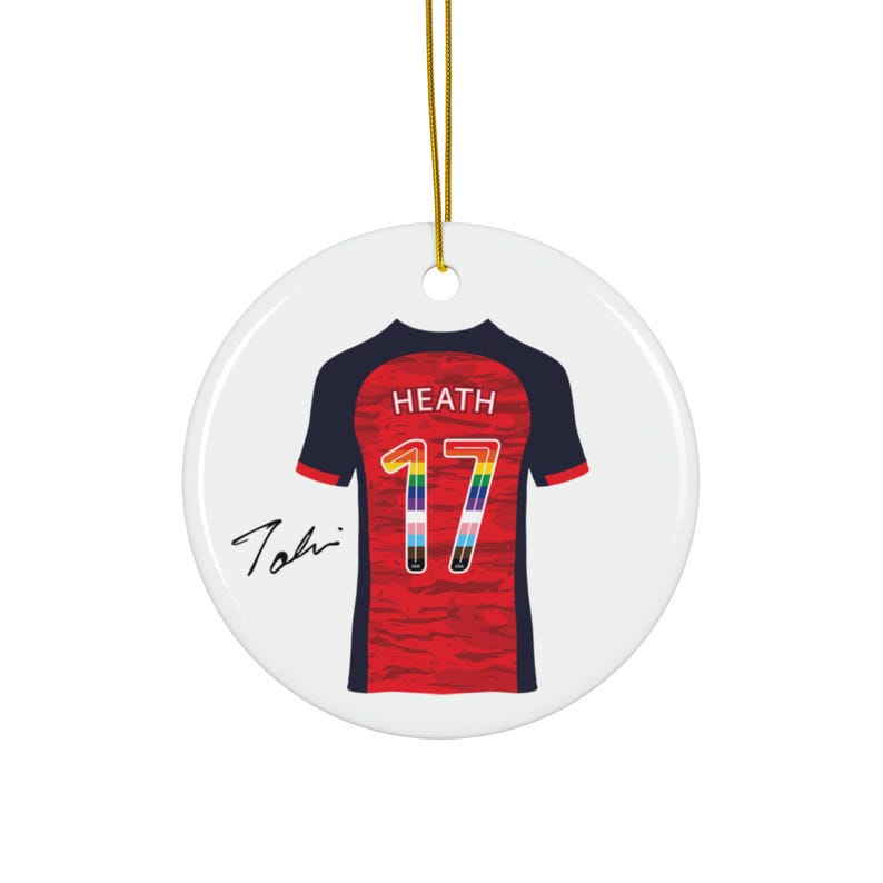 Op de afbeelding: Een ronde, witte keramische ornament met een voetbalshirtontwerp. Het rode shirt heeft marineblauwe mouwen en het regenbooggekleurde nummer 17. De naam "HEATH" staat boven het nummer, met een handtekening links.
