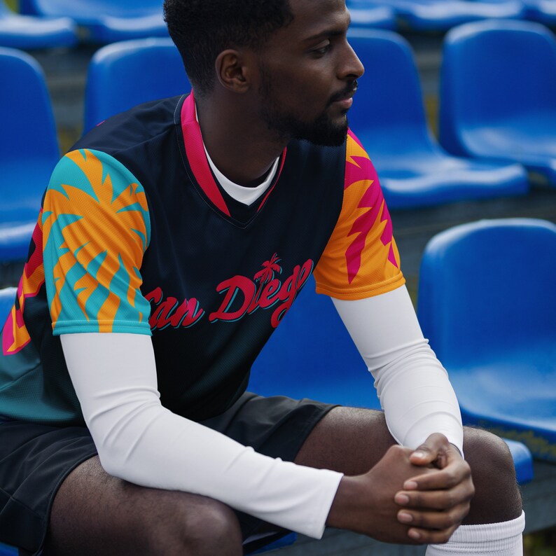 May include: A person wearing a black jersey with a colorful palm tree design and the text "San Diego" on the front. The jersey has white sleeves and the person is sitting on a blue bleacher.