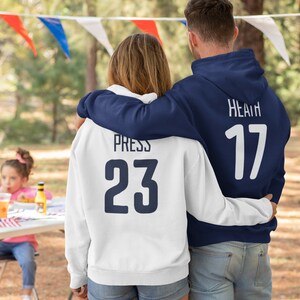 Może przedstawiać: Para w dopasowanych, granatowo-białych bluzach z kapturem z nadrukowanymi imionami i numerami na plecach. Bluza kobiety ma napis "Press 23", a mężczyzny "Heath 17".