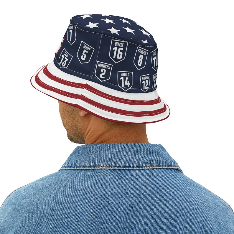 May include: A blue and white bucket hat with an American flag design. The hat has a navy blue crown with white stars and a red, white, and blue brim. The crown features a series of white numbers and names in navy blue shields.