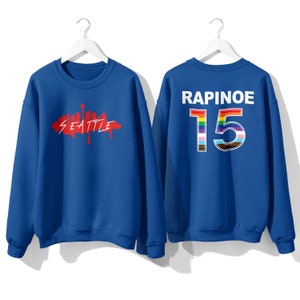 May include: Two royal blue sweatshirts. One has a red graphic of the Seattle skyline with the word "Seattle" underneath. The other has the word "RAPINOE" and the number "15" in rainbow colors.