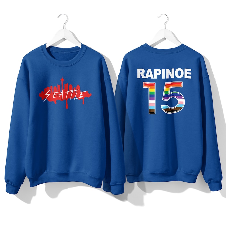 May include: Two royal blue sweatshirts. One has a red graphic of the Seattle skyline with the word "Seattle" underneath. The other has the word "RAPINOE" and the number "15" in rainbow colors.