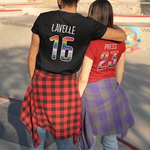 K&ouml;nnte beinhalten: Zwei Personen tragen passende T-Shirts mit Regenbogen-Nummern auf dem R&uuml;cken. Die Person links tr&auml;gt ein schwarzes T-Shirt mit dem Namen "Lavelle" und der Nummer "16". Die Person rechts tr&auml;gt ein rotes T-Shirt mit dem Namen "Press" und der Nummer "23".