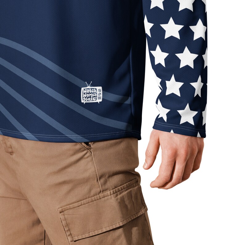 May include: A navy blue long-sleeved shirt with a white star pattern on the right sleeve. The shirt has a white graphic on the front that says "Watch Women Score!" with a television icon.