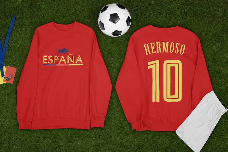 May include: Red long-sleeve sweatshirt with the text "ESPA&Ntilde;A" and a bull graphic on the front and the text "HERMOSO 10" on the back. A white soccer ball and a white pair of sweatpants are also in the image.