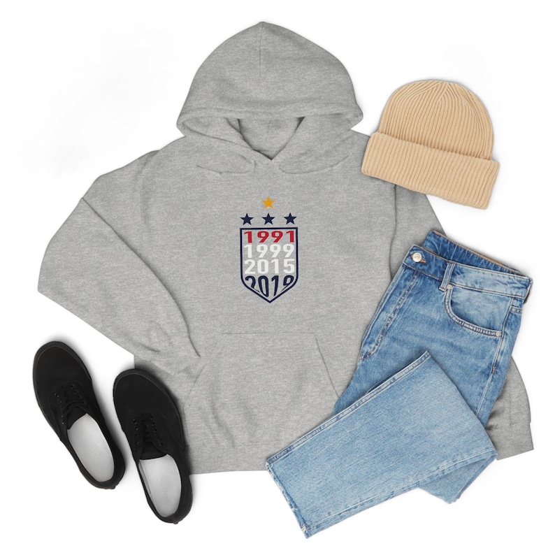 May include: A grey hoodie with a white shield featuring the years 1991, 1999, 2015, and 2019. The shield has three stars above the years and is surrounded by a red, white and blue border. The hoodie is laid out on a white surface with a pair of black trainers, a pair of blue jeans and a beige beanie.