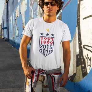 Puede incluir: Una camiseta blanca con un gr&aacute;fico de escudo azul y rojo con los a&ntilde;os 1991, 1999, 2015 y 2019.