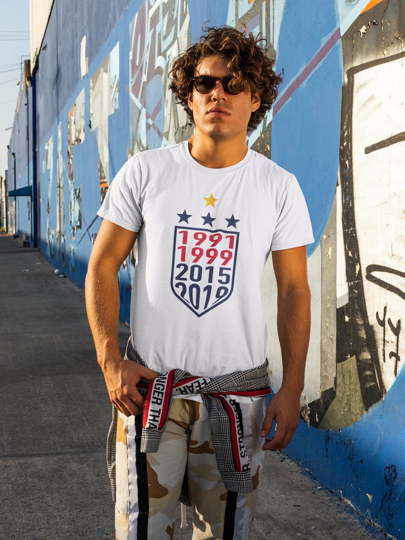 Puede incluir: Una camiseta blanca con un gr&aacute;fico de escudo azul y rojo con los a&ntilde;os 1991, 1999, 2015 y 2019.