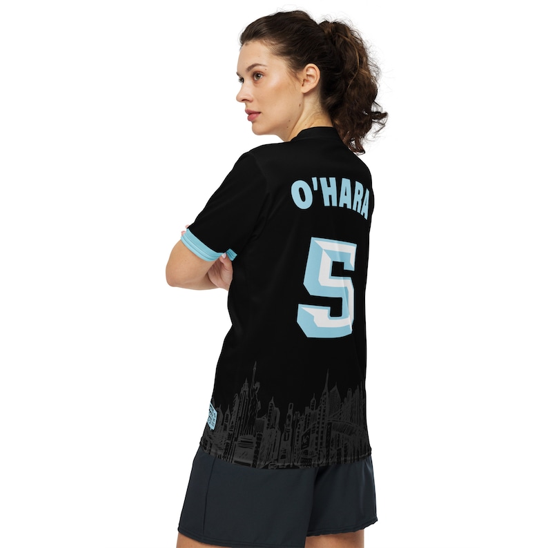 May include: Black soccer jersey with a light blue city skyline design on the bottom. The back of the jersey has the name "O'HARA" and the number "5" in light blue.