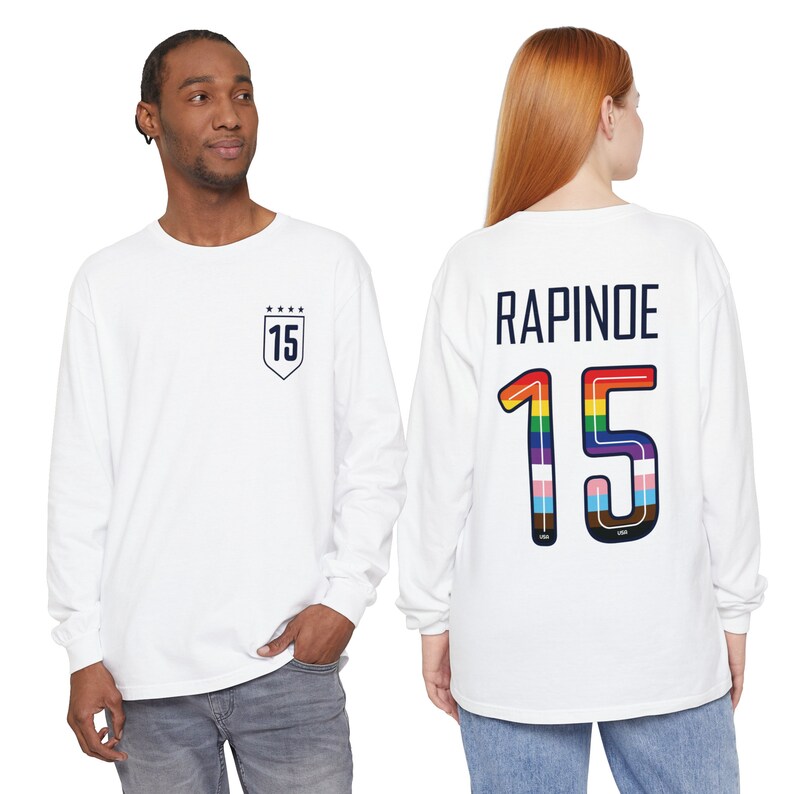 Op de afbeelding: Witte longsleeve t-shirt met een regenboogkleurig nummer "15" op de rug en de naam "RAPINOE" erboven. De voorkant van het shirt heeft een donkerblauw schild met het nummer "15" en vijf sterren.