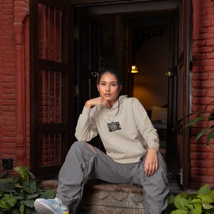 May include: A person wearing a light gray hoodie with a black and white graphic of a television screen with the text "MEGA SUPER SMASH BROS" on it. The person is sitting on a set of stairs with their legs crossed. They are wearing gray pants and colorful sneakers.