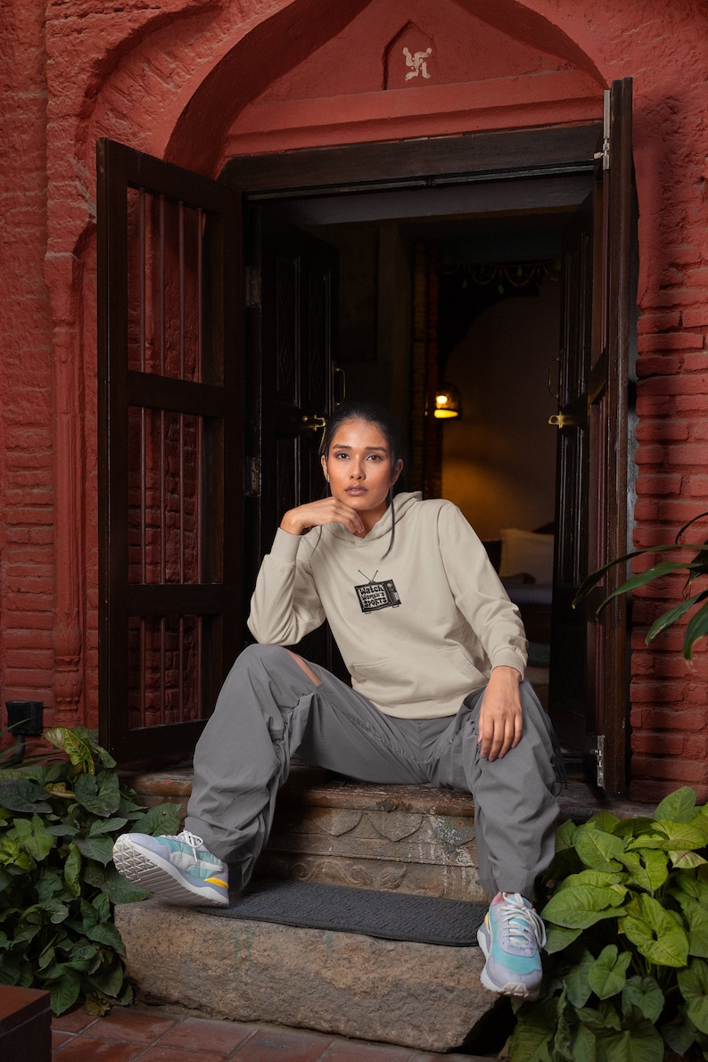 May include: A person wearing a light gray hoodie with a black and white graphic of a television screen with the text "MEGA SUPER SMASH BROS" on it. The person is sitting on a set of stairs with their legs crossed. They are wearing gray pants and colorful sneakers.