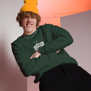 May include: A person wearing a dark green sweatshirt with a white graphic of a television with the text "Watch's" inside. The person is wearing a yellow beanie and black trousers.