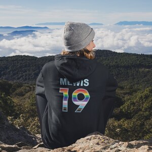 Puede incluir: Una persona lleva una sudadera con capucha negra con el texto "MEWIS 19" en la espalda. El n&uacute;mero 19 est&aacute; rodeado de colores arco&iacute;ris. La persona est&aacute; de pie en un acantilado rocoso con vistas a una cordillera con nubes en el fondo.