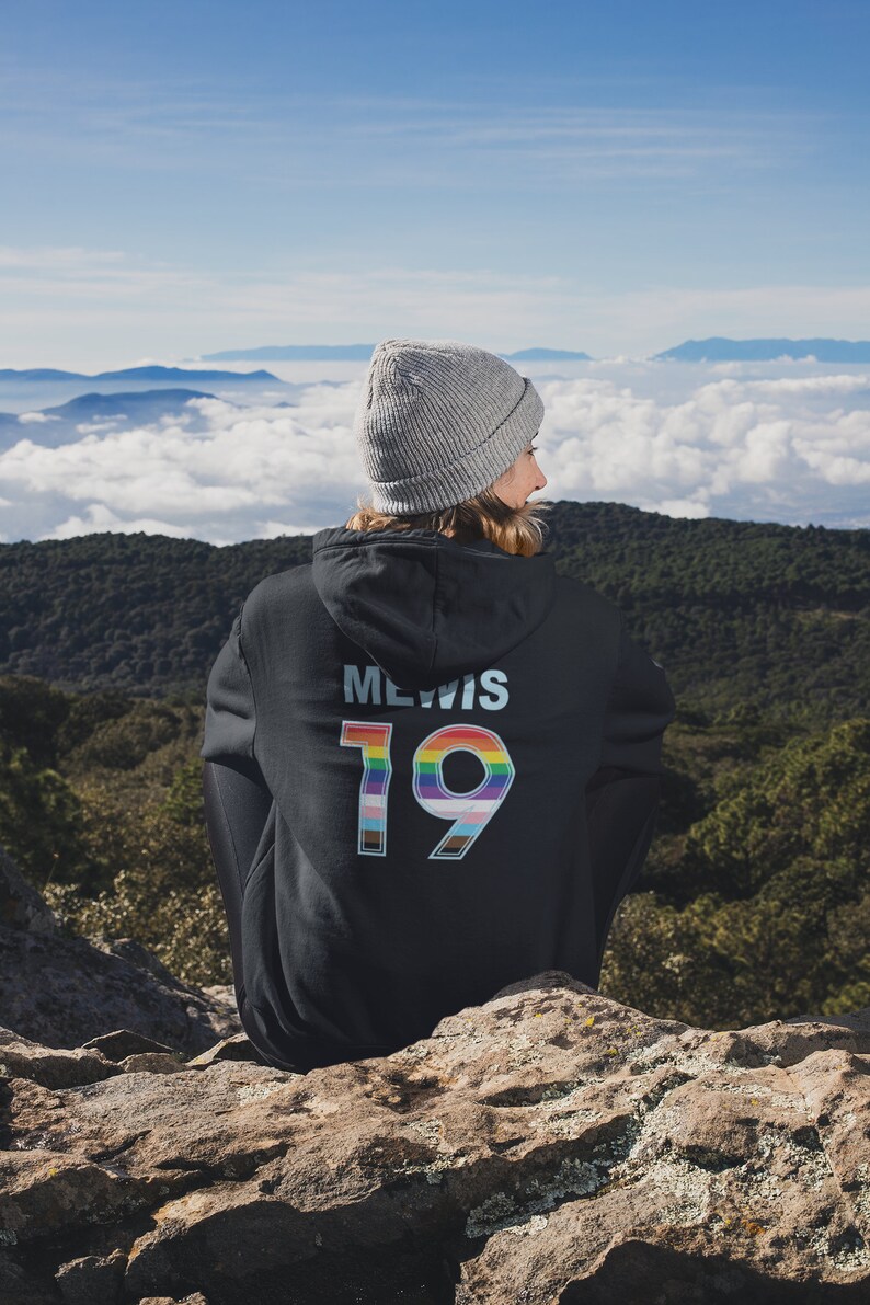 Puede incluir: Una persona lleva una sudadera con capucha negra con el texto "MEWIS 19" en la espalda. El n&uacute;mero 19 est&aacute; rodeado de colores arco&iacute;ris. La persona est&aacute; de pie en un acantilado rocoso con vistas a una cordillera con nubes en el fondo.