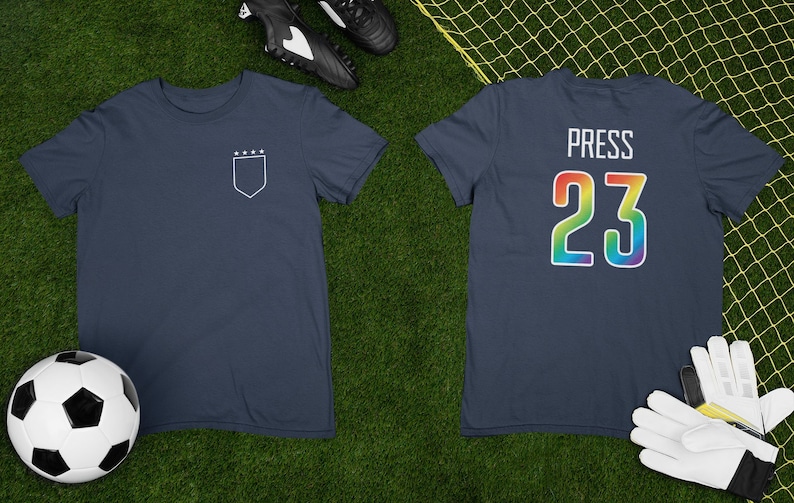 May include: A navy blue t-shirt with a white outline of a shield with five stars on the left chest and the text "PRESS 23" in rainbow colors on the back.