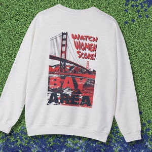 May include: A light gray sweatshirt with a graphic print of the Golden Gate Bridge and the text "Watch Women Score! Bay Area".