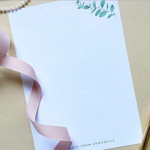 May include: A white stationery notecard with a green leafy branch design in the top right corner. The text "NOTE FROM ANNABELLE" is printed in black at the bottom of the card.