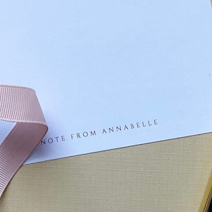 May include: A white card with the text "NOTE FROM ANNABELLE" printed on the bottom. A pink ribbon and a gold pen are on the card.