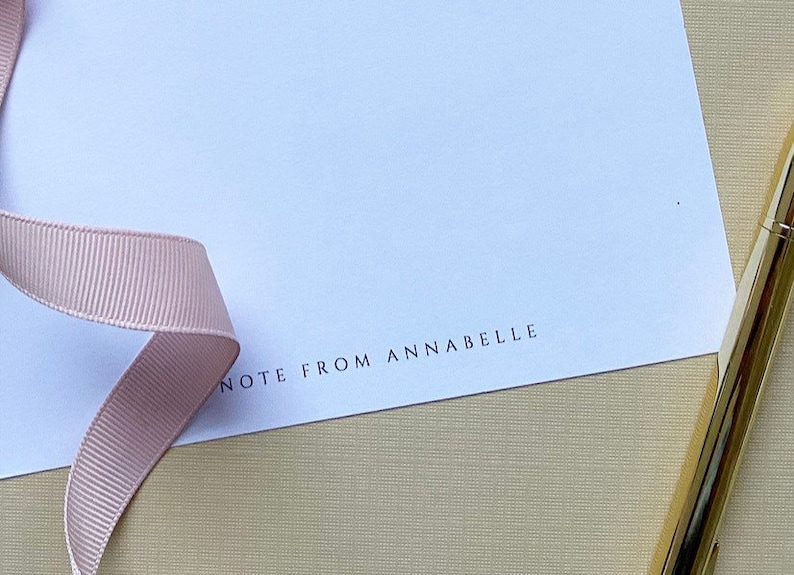 May include: A white card with the text "NOTE FROM ANNABELLE" printed on the bottom. A pink ribbon and a gold pen are on the card.