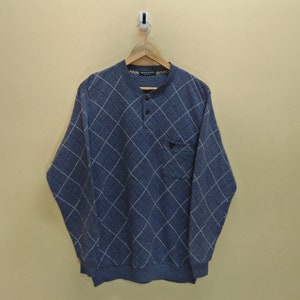 May include: A blue long-sleeved shirt with a white diamond pattern and a single button closure at the neck. The shirt has a small pocket on the left side.