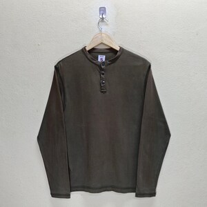 May include: A long-sleeved, olive-green Henley shirt with a button-up placket. The shirt is hanging on a wooden hanger against a neutral background. The shirt has a small, circular tag near the collar.