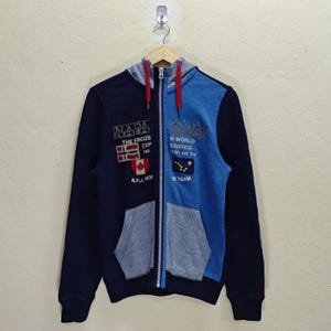 May include: A navy and light blue zip-up hoodie with a grey hood and pocket. The hoodie features patches with text and a Canadian flag. The text includes "NAPA", "THE FROZE EXP", "N WORLD ERIENCE", and "GE TEAM".