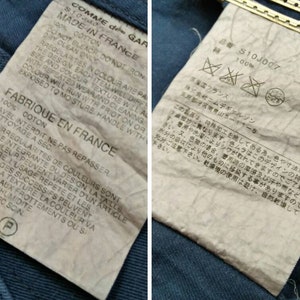 May include: A white tag with black text detailing the care instructions for a garment made of 100% cotton. The tag reads "COMME des GARCONS" and "MADE IN FRANCE". The tag also includes instructions for washing, drying, and ironing the garment. The tag is attached to a blue denim fabric.