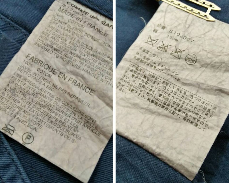 May include: A white tag with black text detailing the care instructions for a garment made of 100% cotton. The tag reads "COMME des GARCONS" and "MADE IN FRANCE". The tag also includes instructions for washing, drying, and ironing the garment. The tag is attached to a blue denim fabric.