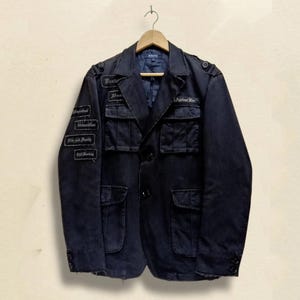 Könnte beinhalten: Dunkelmarineblaue Jacke mit mehreren Taschen und Aufnähern. Die Jacke hat ein klassisches Design mit Knopfleiste und hängt an einem Holzbügel. Aufnäher mit Text sind auf den Ärmeln und der Brust zu sehen.