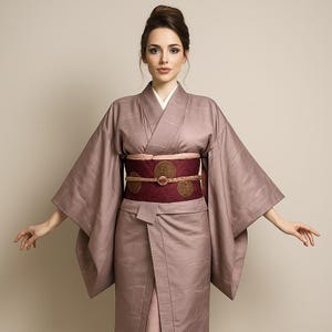 Può includere: Una donna indossa un kimono color malva con colletto bianco e maniche ampie. Una cintura obi bordeaux con disegni circolari dorati stringe la vita. Il kimono è un capo tradizionale giapponese, spesso indossato per occasioni speciali. L'outfit è elegante.