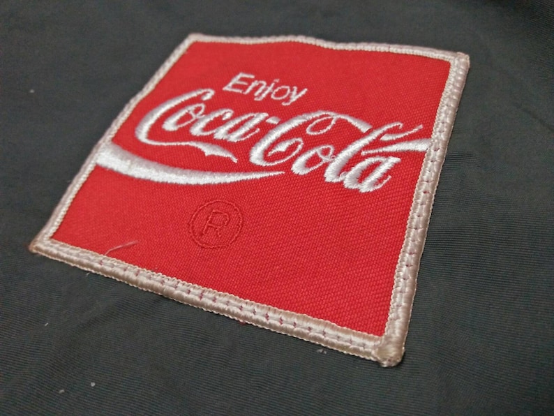 Może przedstawiać: Czerwona naszywka z tkaniny z białym haftem z napisem "Enjoy Coca-Cola" i symbolem znaku towarowego.