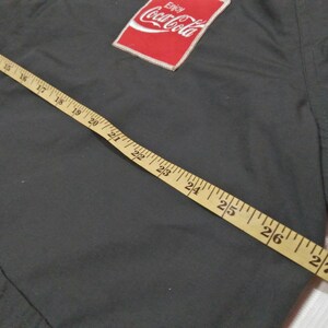 Może przedstawiać: Zielona kurtka z czerwoną naszywką Coca-Cola na lewej piersi. Na kurtce leży miara, kt&oacute;ra wskazuje 71 cm.