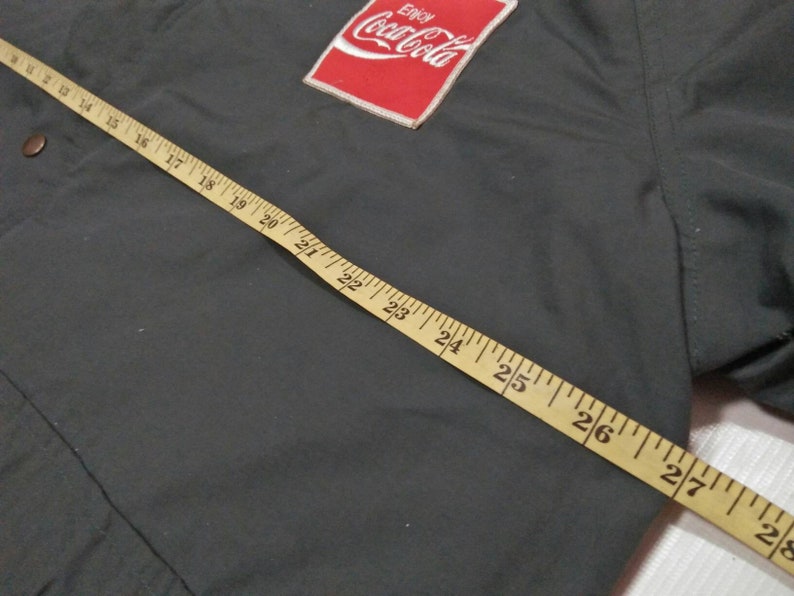 Może przedstawiać: Zielona kurtka z czerwoną naszywką Coca-Cola na lewej piersi. Na kurtce leży miara, kt&oacute;ra wskazuje 71 cm.